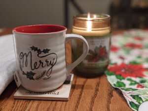 close up of a christmas mug that says "merry" with a candle burning in the background and fabric on a wooden table